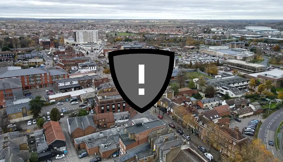 Aerial view of a town with various buildings and streets. A large gray shield with an exclamation mark is overlaid on the image. Cloudy sky.