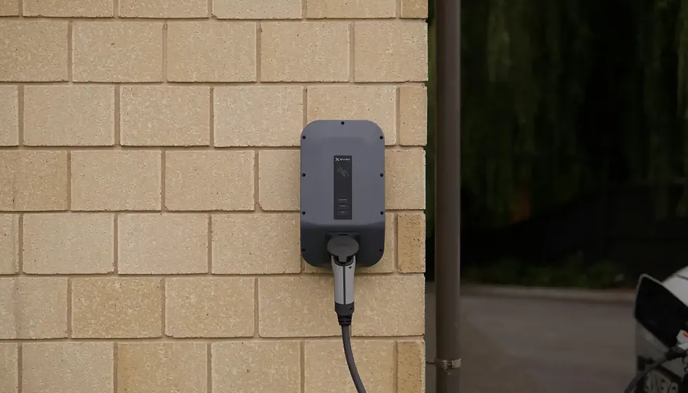 Electric vehicle charger mounted on a beige brick wall. A cable extends downwards. Nearby is a dimly lit outdoor area with greenery.