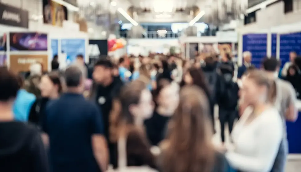 Blurred image of a busy crowd at an indoor event, with colorful posters in the background. People are chatting and walking around.