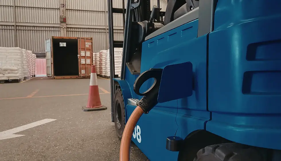 Blue electric forklift charging, orange cable attached. Warehouse setting with pallets, open container, and safety cone visible.